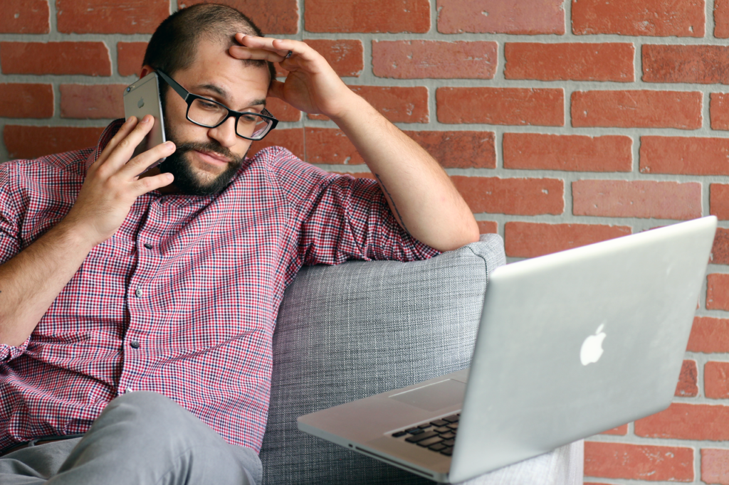 5 señales de que tu web está frenando el crecimiento de tu empresa (aunque no lo parezca) 1 Un hombre con barba parece frustrado mientras trabaja a distancia con su portátil y su teléfono.
